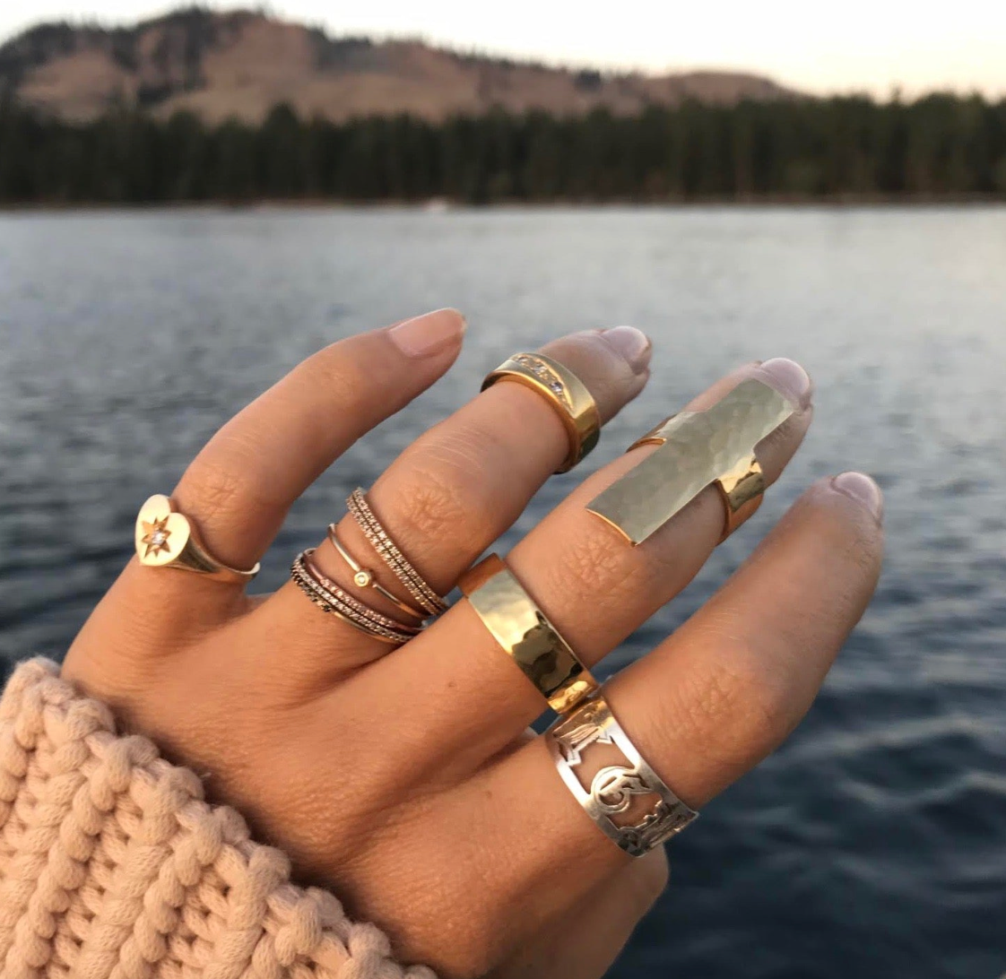Hand wearing multiple gold rings with a blurred lake and trees in the background. On the pinky finger is the charlotte heart starburst ring. 