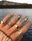 Hand wearing multiple gold rings with a blurred lake and trees in the background. On the pinky finger is the charlotte heart starburst ring. 