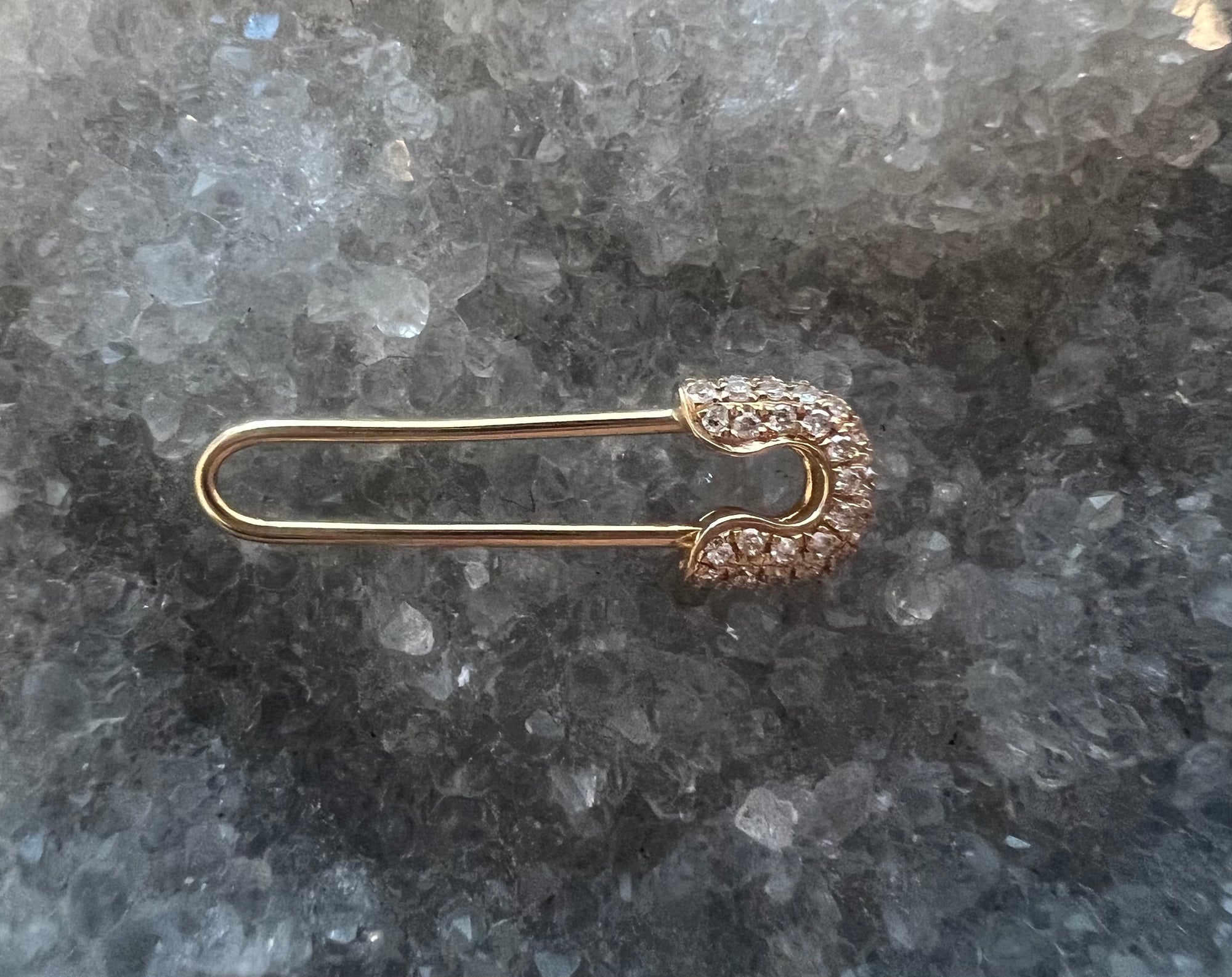 A gold safety pin brooch or earring with a pavé-set crystal crescent-shaped head, displayed on a textured gray and white crystal background.