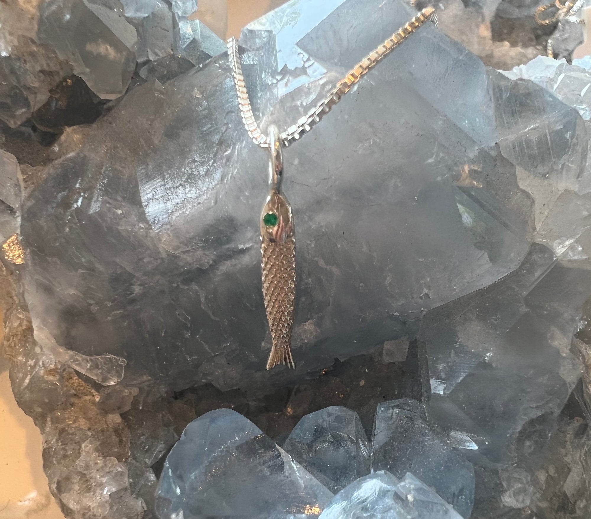 Handmade silver-fish and sardine pendant with green emerald gemstone eye on a silver box-chain, placed on a crystal rock.