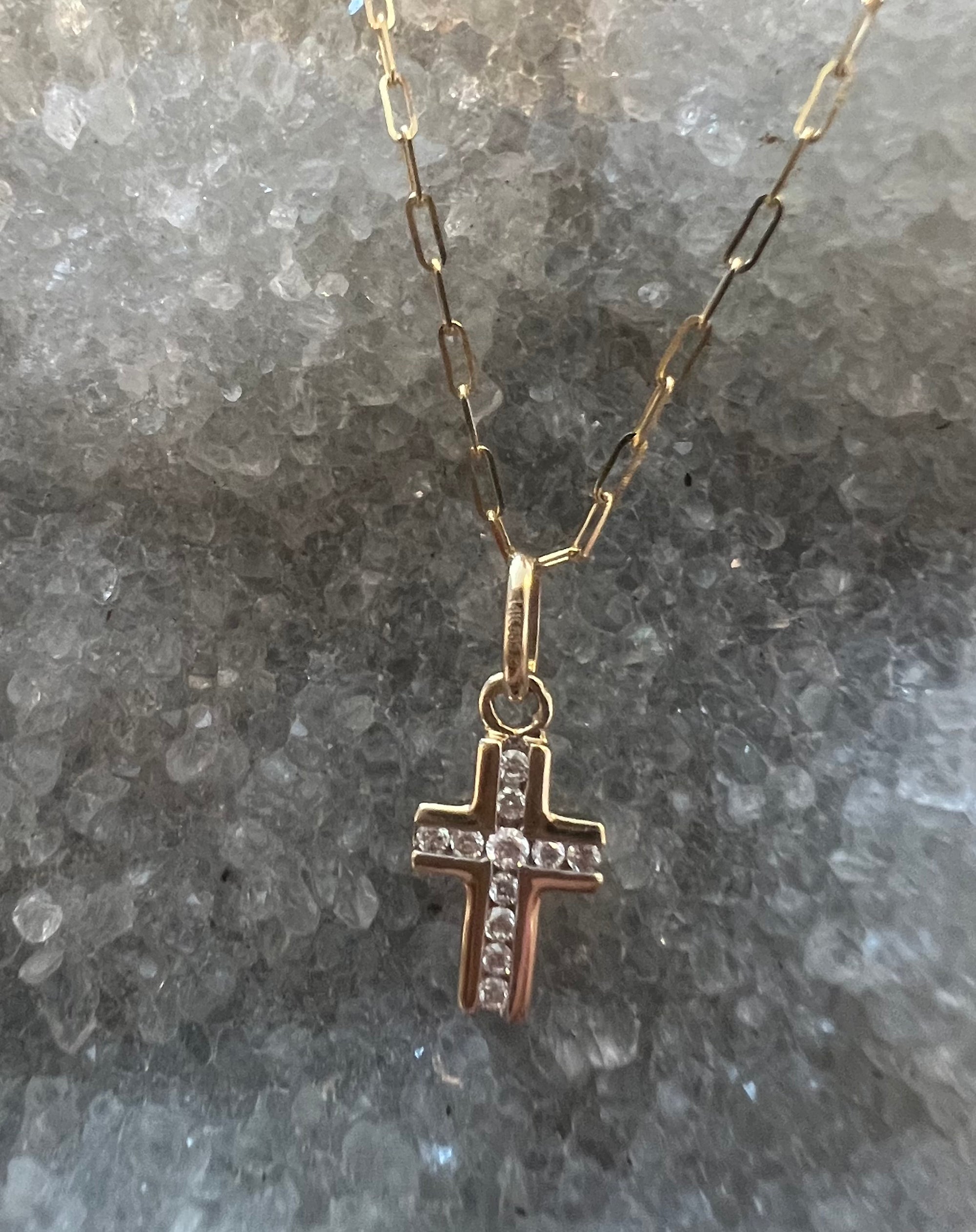 A solid 14k gold cross pendant necklace with natural VS1 diamonds inlaid in the center, resting on a textured gray crystal background.