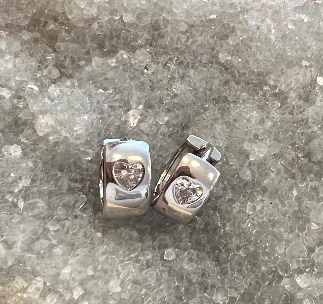 A pair of polished sterling silver huggie earrings with a heart-shaped clear cubic zarconia gemstones, displayed on a textured gray and white crystalline surface.