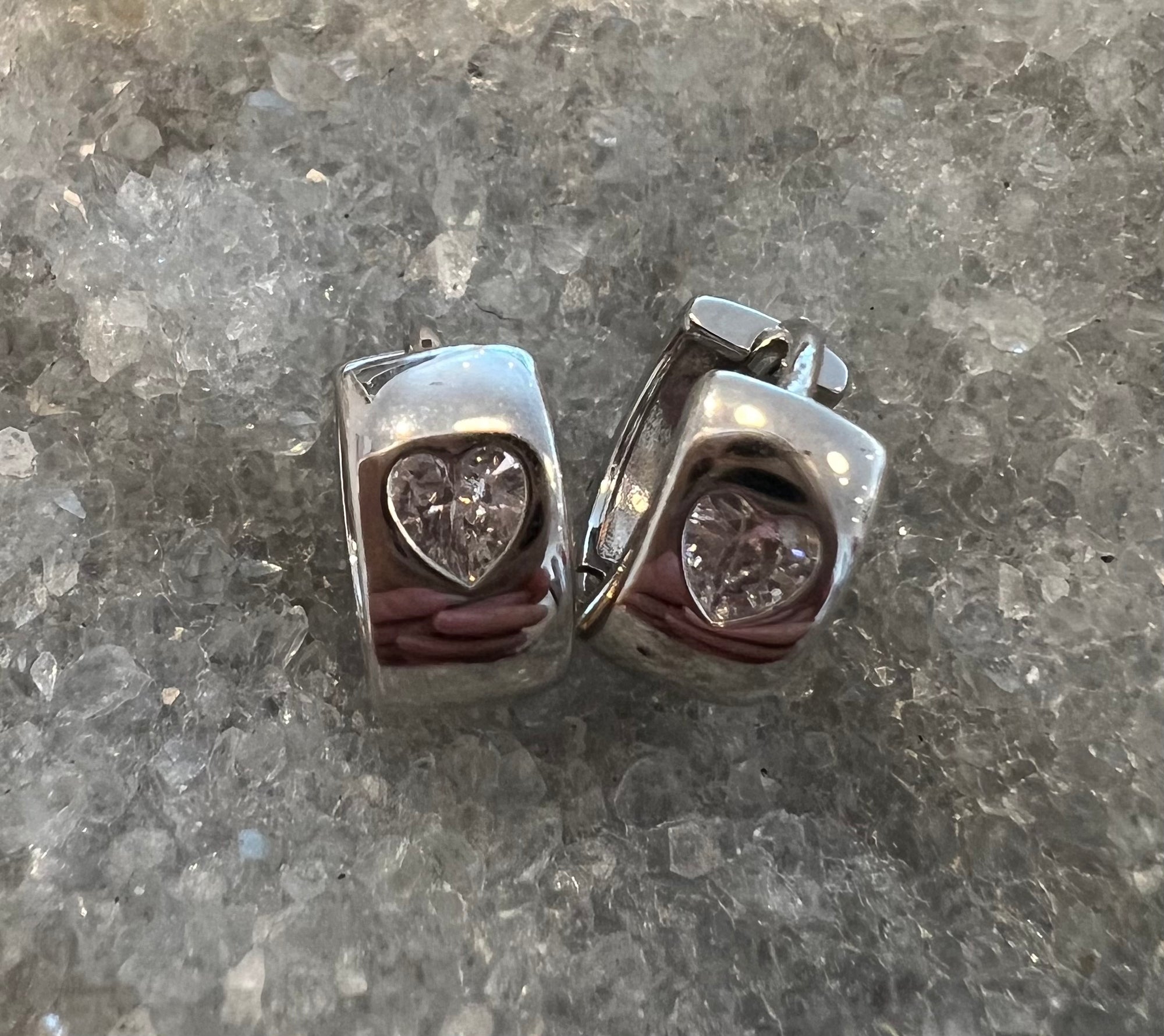 A pair of sterling silver hoop earrings with heart-shaped clear crystal cubic zarconia, displayed on a textured gray stone surface.