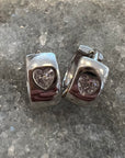 A pair of sterling silver hoop earrings with heart-shaped clear crystal cubic zarconia, displayed on a textured gray stone surface.