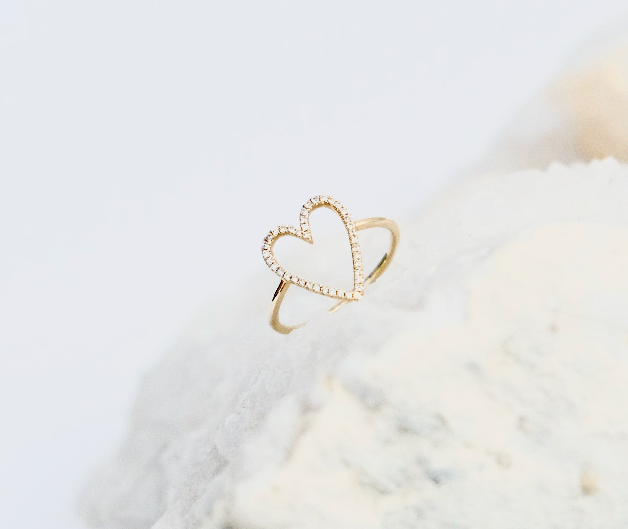 A solid 14k gold heart-shaped halo ring with small diamond gemstones along the outline, displayed on a textured white surface against a soft white background.