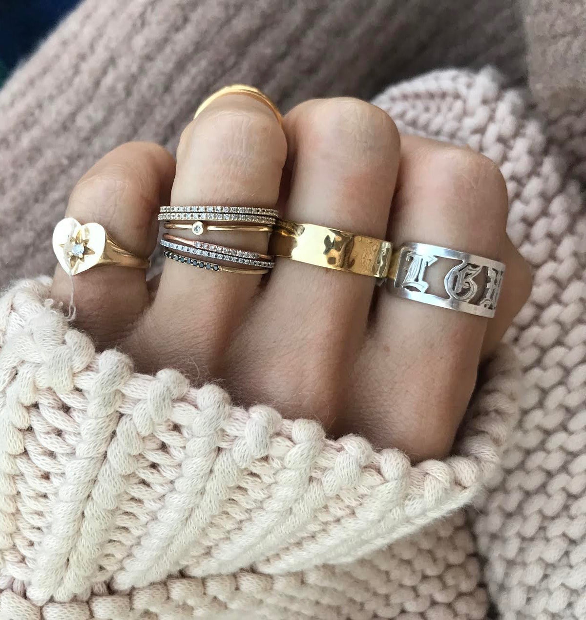 A hand showcasing many different rings in silver and gold vermeil. With a hammered textured cigar band on the middle finger, with a sweater texture in the background. 