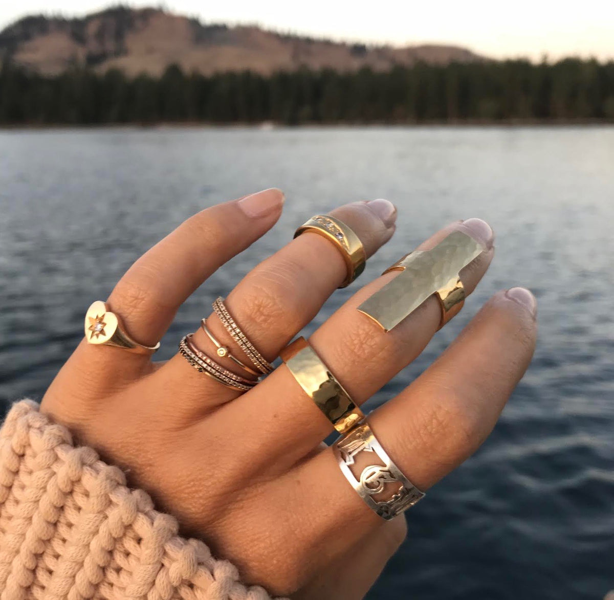 A hand wearing multiple JNB Style rings, with a Stella Ring, a hammered cigar band in gold vermeil, on the middle finger. With a lake and mountains in the background. 